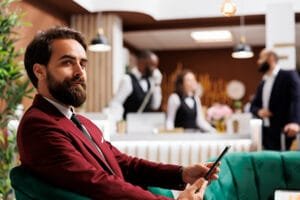 A confident hotel manager sitting in a modern hotel lobby using a smartphone, representing successful SEO for hotels to improve bookings and visibility.